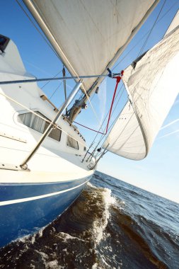 Açık bir yaz gününde durgun bir suda mavi bir yat yüzüyor. Güverteden pruvaya, direğe ve yelkenlere yakın görüntü. Dalgalar ve su sıçramaları. Ruhnu Adası, Estonya, Baltık Denizi