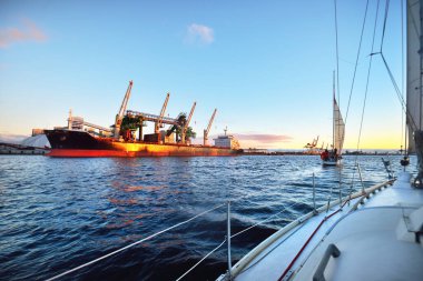 Gün batımında Daugava nehrinde beyaz bir yat. Güverteden pruvaya, direğe ve yelkenlere yakın görüntü. Arka planda yük gemileri ve iskele vinçleri var. Riga Körfezi, Letonya
