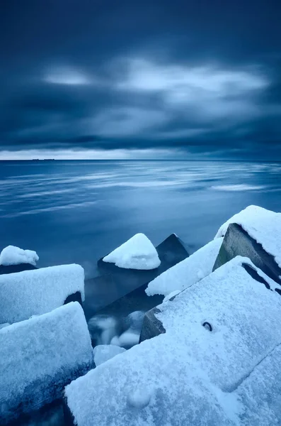 Snow-covered breakwaters close-up, frozen Baltic sea in the background. Dark storm clouds. Winter, seasons, climate change, global warming concepts. Long exposure