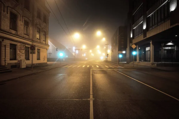 An empty illuminated asphalt road through the old historical buildings and houses in a fog at night. Street lights (lanterns) close-up. Riga, Latvia. Dark cityscape