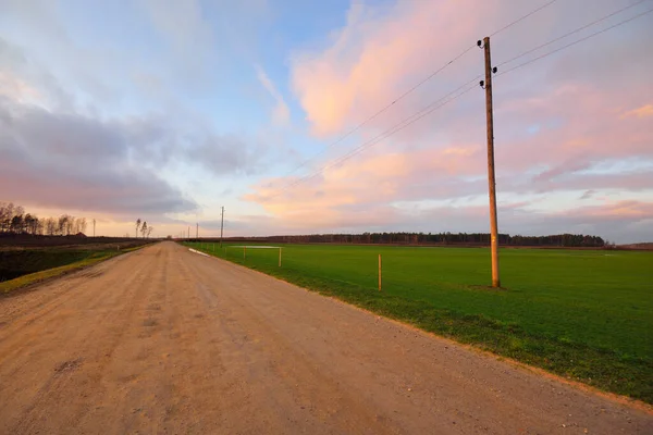 緑豊かな田園農業を通る未舗装の道路は日没時に畑を耕した トランスポールの開閉 美しいピンクの雲の風景 暖かい冬だ 気候変動のテーマ ラトビア — ストック写真