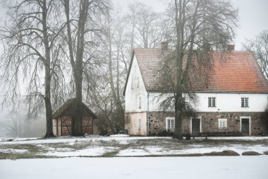Kırmızı kiremitli ve eski yosunlu ağaçlı geleneksel kır evi. Bulutlu bir günde kar fırtınası. Kırsal kesimde kış manzarası. Almanya