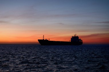 Büyük bir kargo gemisi gün batımında deniz fenerine doğru yola çıkıyor. Renkli bulutlu bir manzara. Riga Körfezi, Baltık Denizi, Letonya. Uluslararası iletişim, lojistik tema