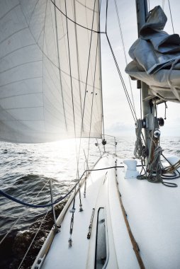 Güneşli bir yaz gününde açık denizde yelken açan beyaz bir yat. Güverteden pruva ve yelkenlere kadar yakın çekim. Dalgalar ve su sıçramaları. Sirus bulutları ve uçak izleri olan açık mavi bir gökyüzü. Hollanda