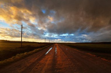 Gün batımında yağmurdan sonra tarlalardan geçen boş bir köy yolu. Dramatik renkli gökyüzü, destansı bulutlar. Transformatör direkleri yakın çekim. Letonya