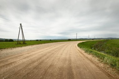 Yağmurdan sonra tarlalardan geçen boş bir köy yolu. Dramatik gökyüzü, destansı bulutlar. Kötü hava. Transformatör direkleri yakın çekim. Letonya