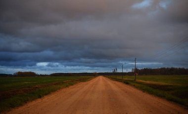Gün batımında yağmurdan sonra tarlalardan geçen boş bir köy yolu. Dramatik renkli gökyüzü, destansı bulutlar. Transformatör direkleri yakın çekim. Letonya