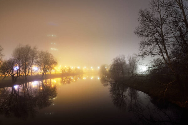 River and a city park in a fog at night. Bright illumination by the street lanterns. Riga, Latvia. Dark cityscape