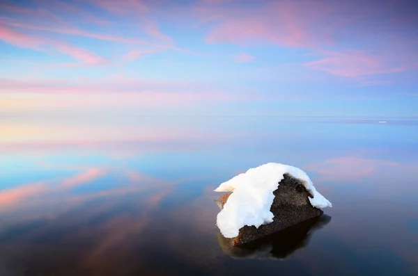 Snow-covered stone close-up, frozen Baltic sea in the background. Latvia. Colorful pink sunset clouds, symmetry reflections on the water. Winter, seasons, climate change, global warming concepts