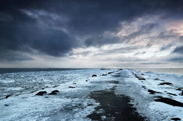 Empty promenade and snow-covered breakwaters close-up, frozen Baltic sea in the background. Dark storm clouds. Winter, seasons, climate change, global warming concepts. Long exposure