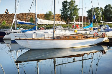 Açık bir yaz gününde, yakınlarda beyaz ahşap bir yat iskeleye demirlemiş. Arka planda yeşil ağaçlar ve yakacak odun var. Riga Limanı, Letonya