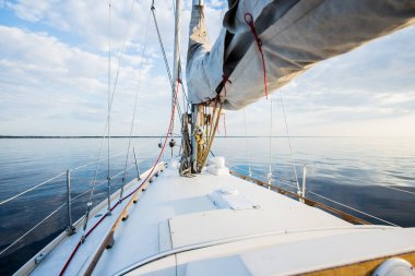 Gün batımında durgun suda yüzen beyaz bir yat. Güverteden pruvaya, direğe ve yelkenlere bir manzara. Şaşırtıcı bulut manzarası. Doğal ayna. Spor ve eğlence teması. Baltık Denizi, Letonya