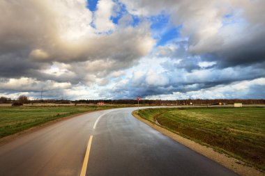 Sonbahar. Boş asfalt yolda fırtına bulutları. Arka planda tarım alanları ve orman var. Sıcak akşam güneşi. Baltık Denizi, Garciems, Letonya