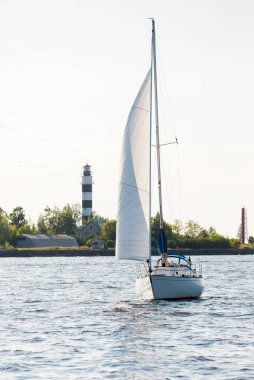 Açık bir günde beyaz yelkenli tekneyle denize açılmak. Arka planda deniz feneri ve orman var. Riga Körfezi, Baltık Denizi, Letonya