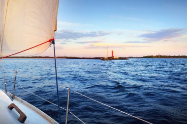 Günbatımında beyaz yat, arka planda kırmızı deniz feneri. Güverteden pruvaya ve yelkenlere bir manzara. Şaşırtıcı bulut manzarası. Spor ve eğlence teması. Riga Körfezi, Baltık Denizi, Letonya