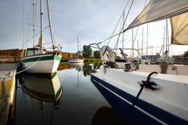 Açık bir yaz gününde açık bir rıhtıma demirlenmiş mavi şalopeli yat, yakın çekim. Arka planda yeşil ağaçlar ve yakacak odun var. Sudaki yansımalar. Riga Limanı, Letonya