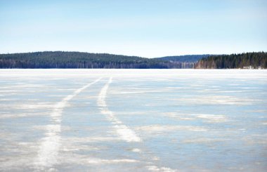 Karla kaplı donmuş nehrin üzerindeki açık mavi gökyüzünde traktör izleri var. Eski yeşil ağaçlar yakın plan. Arka planda kozalaklı orman var. Güneşli kış günü. Karelya Kuzey, Laponya
