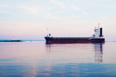 Gün batımında büyük bir kargo gemisi. Renkli akşam bulutları ve parlak güneş ışığı. Arka planda deniz feneri var. Baltık Denizi, Letonya