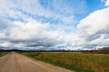 Sonbahar. Boş toprak yolun üzerindeki fırtına bulutları. Arka planda tarım alanları ve orman var. Sıcak akşam güneşi. Baltık Denizi, Garciems, Letonya