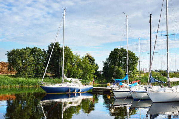 Elegant and modern sailboats (for rent) moored to a pier in a yacht marina on a clear summer day. Riga, Latvia. Symmetry reflections on water. Vacations, sport, amateur recreational sailing, cruise