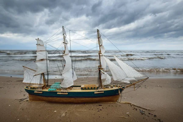 Antiquarian wooden scale model of the clipper tall ship, close-up. Dramatic sky, sandy shore (sand dunes) of the Baltic sea. Traditional craft, souvenir, toy, hobby, vintage, collecting, modeling