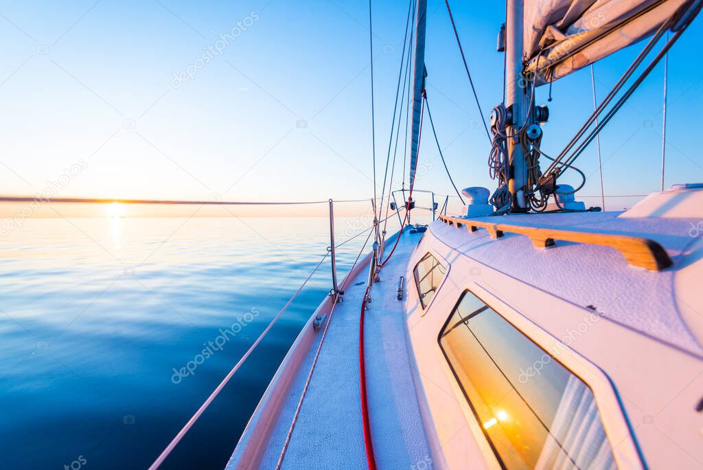 The calm water. White sloop rigged yacht sailing at sunset. A view from the deck to the bow, mast and the sails. Baltic Sea, Latvia