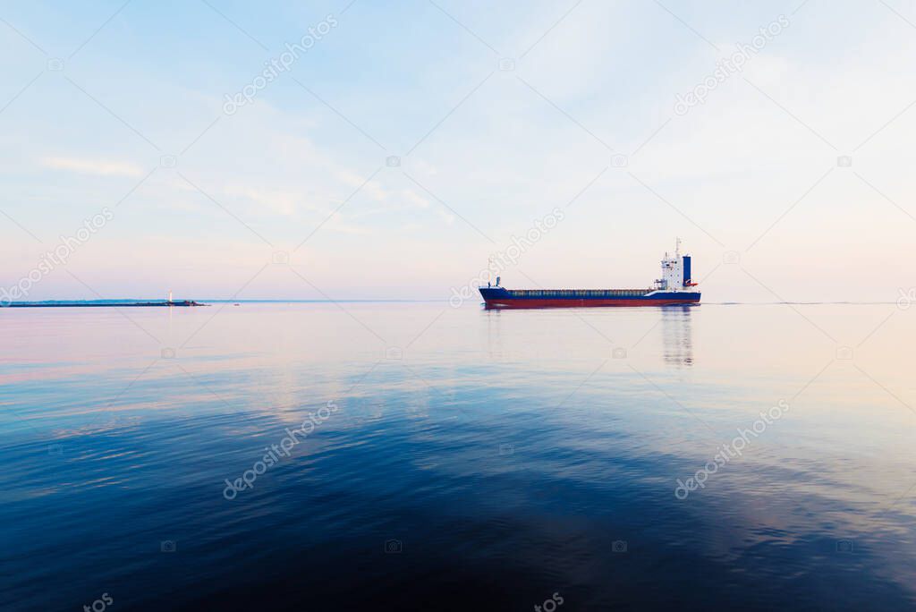 Large cargo ship at sunset. Colorful evening clouds and bright sunlight. lighthouse in the background. Baltic Sea, Latvia