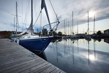 Gün batımında bir rıhtıma demirlenmiş mavi şalopeli yat, yakın plan. Dramatik akşam gökyüzü. Sudaki yansımalar. Riga Limanı, Letonya