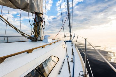 Gün batımında durgun suda yüzen beyaz bir yat. Güverteden pruvaya, direğe ve yelkenlere bir manzara. Şaşırtıcı bulut manzarası. Doğal ayna. Spor ve eğlence teması. Baltık Denizi, Letonya
