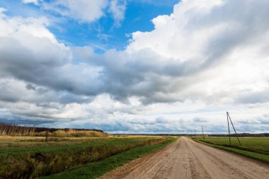 Sonbahar. Boş toprak yolun üzerindeki fırtına bulutları. Arka planda tarım alanları ve orman var. Sıcak akşam güneşi. Baltık Denizi, Garciems, Letonya