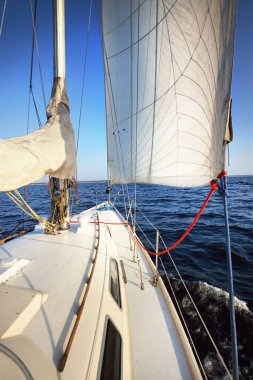Güneşli bir günde beyaz bir yat denize açılıyor. Güverteden pruva ve yelkenlere kadar yakın çekim. Dalgalar ve su sıçramaları. Britanya, Fransa