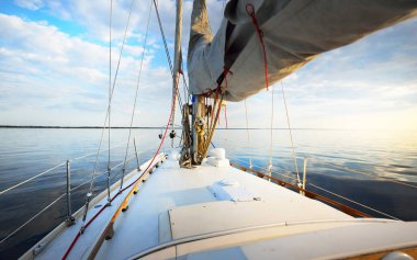 Gün batımında durgun suda yüzen beyaz bir yat. Güverteden pruvaya, direğe ve yelkenlere bir manzara. Şaşırtıcı bulut manzarası. Doğal ayna. Spor ve eğlence teması. Baltık Denizi, Letonya