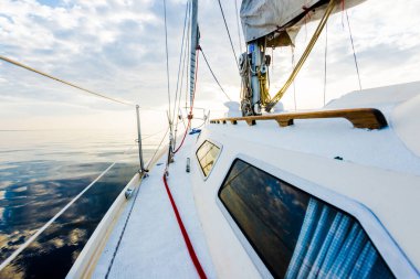 Gün batımında durgun suda yüzen beyaz bir yat. Güverteden pruvaya, direğe ve yelkenlere bir manzara. Şaşırtıcı bulut manzarası. Doğal ayna. Spor ve eğlence teması. Baltık Denizi, Letonya