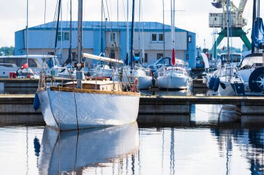 Açık bir yaz gününde, yakınlarda beyaz ahşap bir yat iskeleye demirlemiş. Arka planda tekneler var. Riga Limanı, Letonya