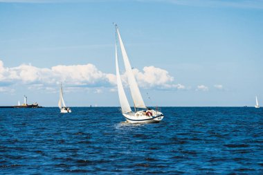Açık bir günde beyaz yelkenli tekneyle denize açılmak. Arka planda deniz feneri var. Bulutlu mavi gökyüzü. Riga Körfezi, Baltık Denizi, Letonya