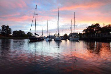 Yat ve tekneler gün batımında bir rıhtıma demirlemiş, yakın plan. Renkli bulutlu dramatik akşam gökyüzü. Sudaki yansımalar. Riga Limanı, Letonya