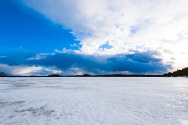Karla kaplı donmuş Kuito gölünün üzerindeki dramatik mavi gökyüzü, arka planda kozalaklı orman. Kış kır manzarası. Fırtına bulutları. Karelya Kuzey, Laponya