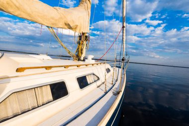 Gün batımında durgun suda yüzen beyaz bir yat. Güverteden pruvaya, direğe ve yelkenlere bir manzara. Şaşırtıcı bulut manzarası. Doğal ayna. Spor ve eğlence teması. Baltık Denizi, Letonya
