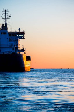 Gün batımında büyük bir kargo gemisi, yakın plan. Arka planda deniz feneri var. Riga, Letonya