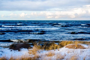 Güneşli bir kış gününde, karla kaplı deniz kıyısında. Kuzey denizi üzerinde soğuk fırtınalı dalgalar ve bulutlar, Hollanda