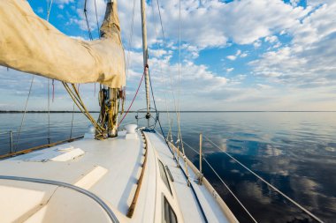 Gün batımında durgun suda yüzen beyaz bir yat. Güverteden pruvaya, direğe ve yelkenlere bir manzara. Şaşırtıcı bulut manzarası. Doğal ayna. Spor ve eğlence teması. Baltık Denizi, Letonya