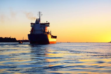 Gün batımında büyük bir kargo gemisi ve bir pilot bot, yakın plan. Arka planda deniz feneri var. Gökyüzü açık. Riga, Letonya