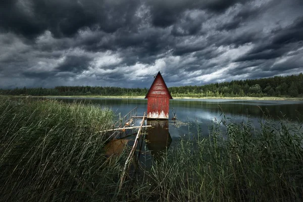 A small red decorative wooden house (log cabin) near the lake and the forest, close-up, Latvia. Dramatic clouds before the thunderstorm. Idyllic rural scene