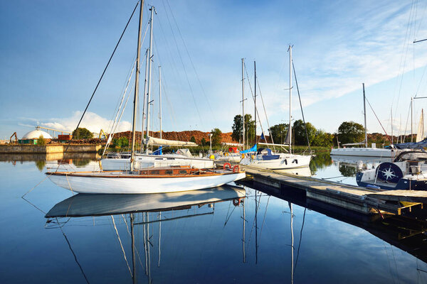 Белая деревянная яхта пришвартована к пирсу в ясный летний день крупным планом. Зеленые деревья и дрова на заднем плане. Рижский порт, Латвия