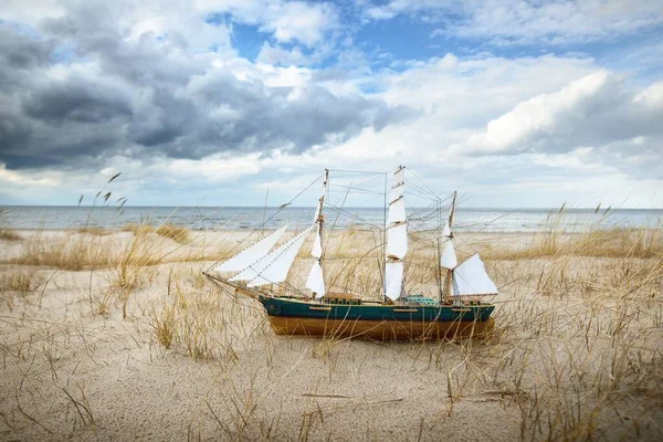 Antiquarian wooden scale model of the clipper tall ship, close-up. Dramatic sky, sandy shore (sand dunes) of the Baltic sea. Traditional craft, souvenir, toy, hobby, vintage, collecting, modeling