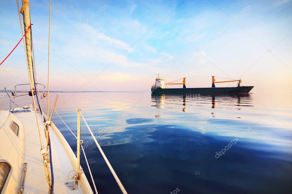 Large cargo crane ship at sunset. Evening clouds and bright sunlight. Baltic Sea, Latvia. View from a sailing boat