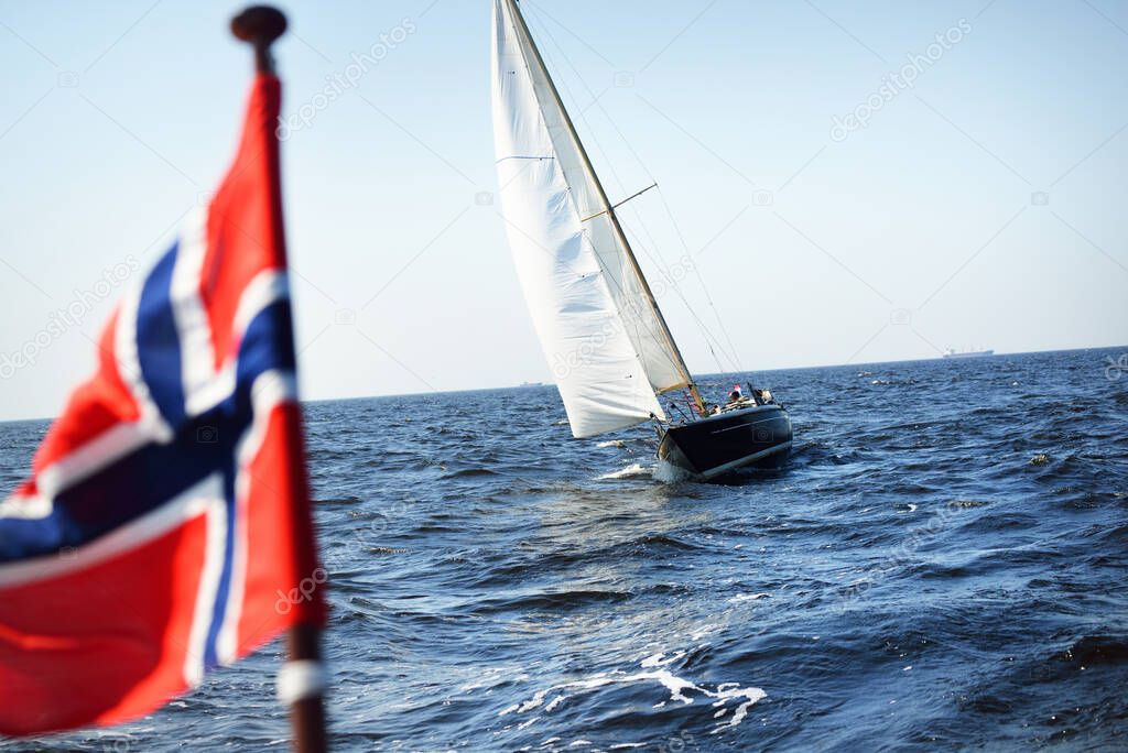 Blue yacht sailing on a sunny summer day. Side view of white sails. Norway flag close-up. Waves and water splashes. Baltic sea, Sweden. Transportation, sport, recreation, regatta, leisure activity