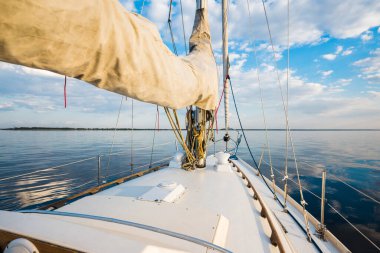 Gün batımında durgun suda yüzen beyaz bir yat. Güverteden pruvaya, direğe ve yelkenlere bir manzara. Şaşırtıcı bulut manzarası. Doğal ayna. Spor ve eğlence teması. Baltık Denizi, Letonya