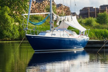 Açık bir yaz gününde, yakın planda mavi bir yat iskeleye demirlemiş. Arka planda yeşil ağaçlar ve yakacak odun var. Riga Limanı, Letonya