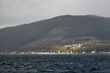 Sudan kayalık nehir kıyılarının panoramik görüntüsü. Arka planda kır evleri, ağaçlar, tepeler ve dağlar var. Bulutlu mavi gökyüzü. Gare Loch, Clyde Firth, İskoçya, İngiltere